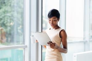 Beautiful african american young businesswoman with cup of coffee holding clipboard and reading in office