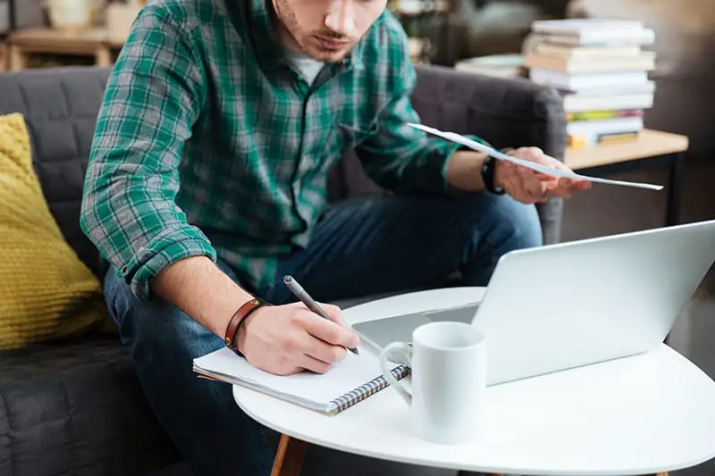 Cropped image of man in green shirt sitting on sofa with laptop and documents and writing by the table. Coworking
