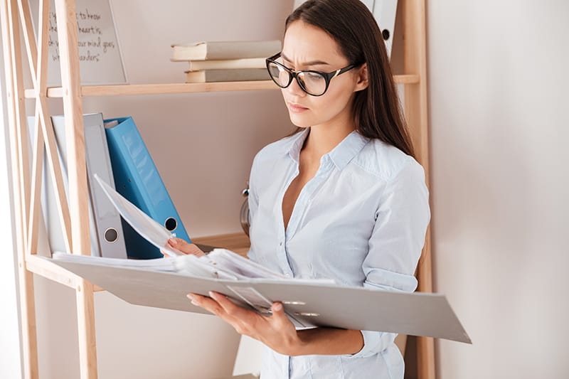 Young concentrated businesswoman holding folder near book shelf while standing in office