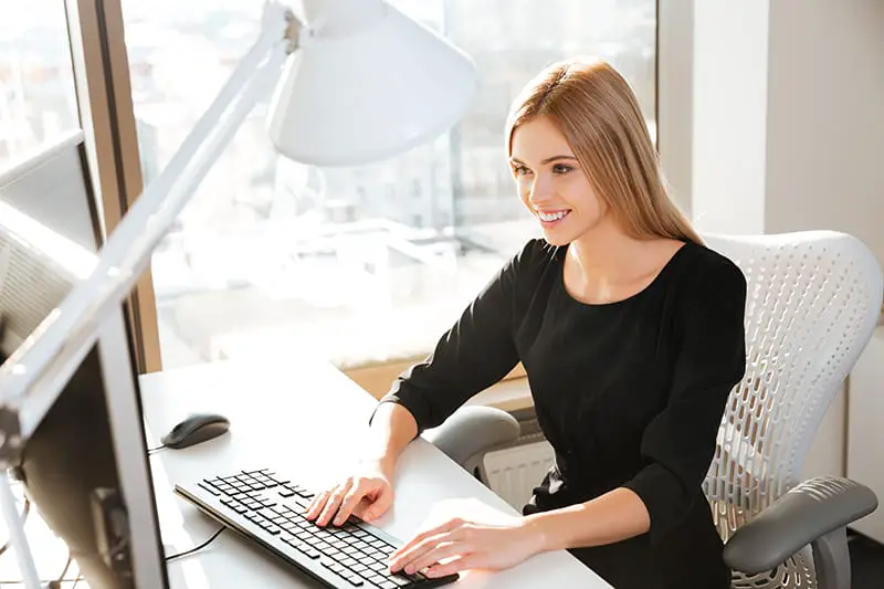 Photo of joyful young lady worker using computer in office and typing by keyboard. Look at computer.