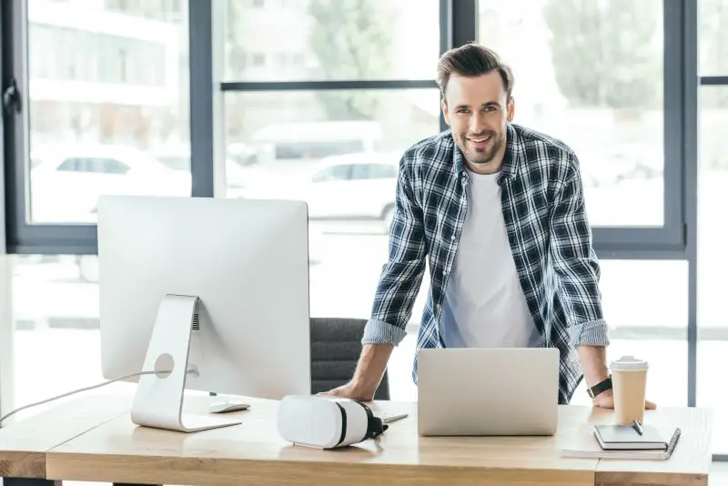 young man smiling at camera while leaning at table with laptop and desktop computer