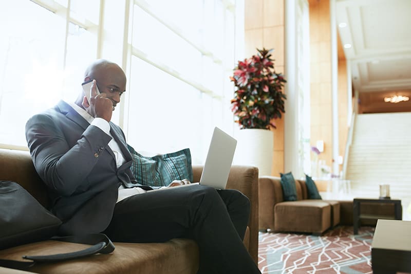 Businessman sitting on sofa working using cell phone and laptop. African male executive waiting in hotel lobby.