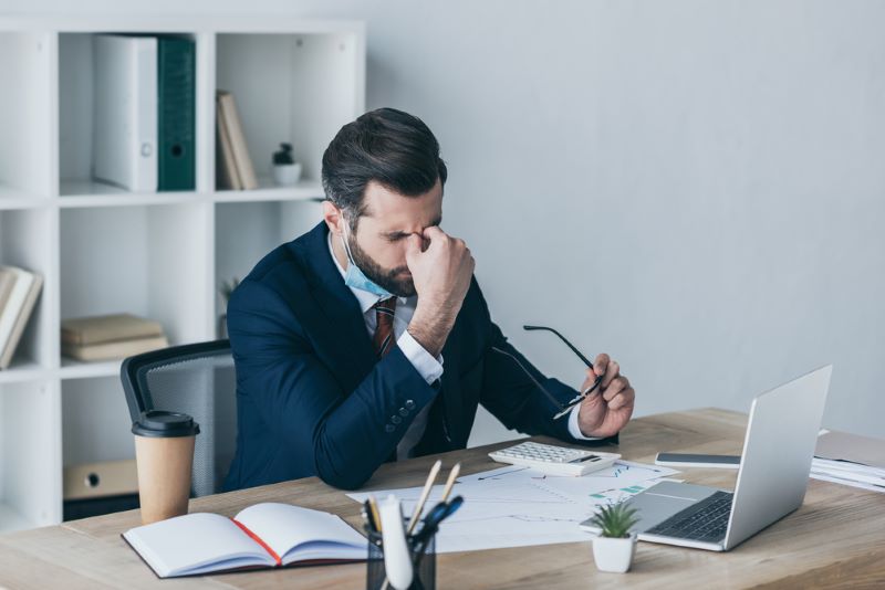 stressed exhausted businessman holding eyeglasses touching face while sitting in workplace