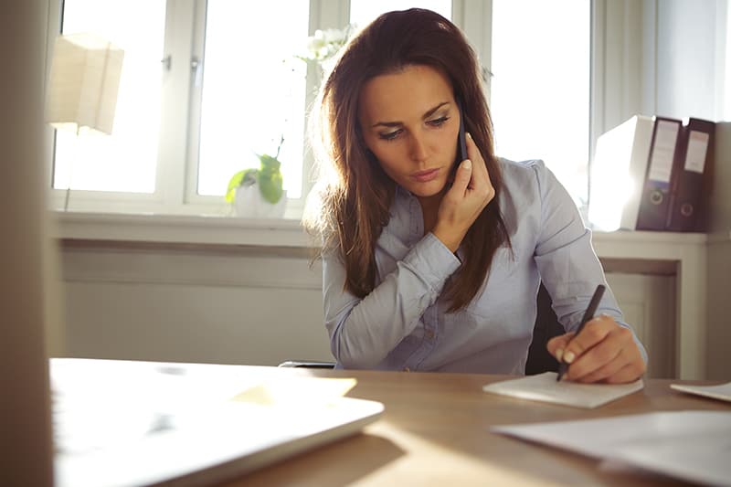 Young woman talking on mobile phone and writing notes while sitting at her desk. Pretty caucasian female working in home office.