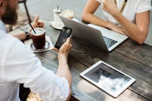 two people sat at a table one lookng at phone the other at laptop