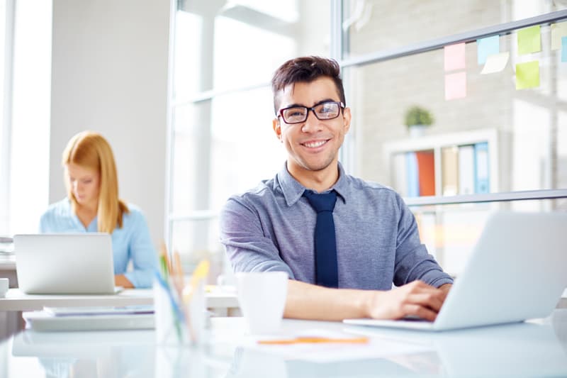 Young man sat at a desk using a laptop, female in the background using laptop