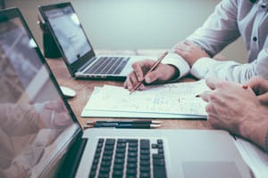 two people sitting side by side looking ad document on table between two laptops