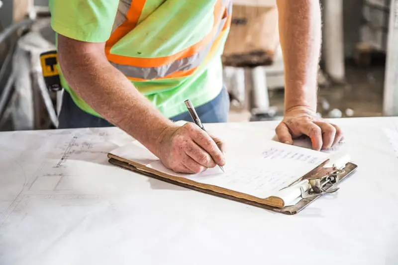 Workman writing on a clipboard placed on top of blueprints