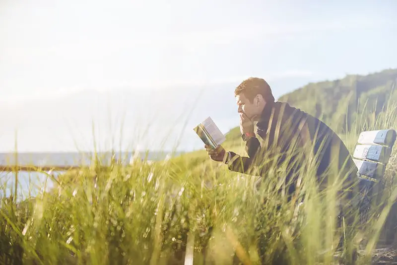 MAn reading book on bench long grass sea view in background
