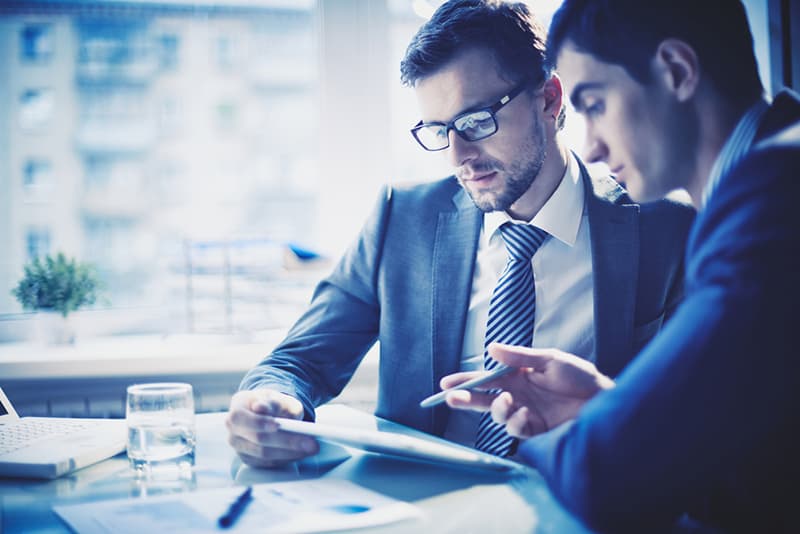 two men sitting at a desk looking at a tablet