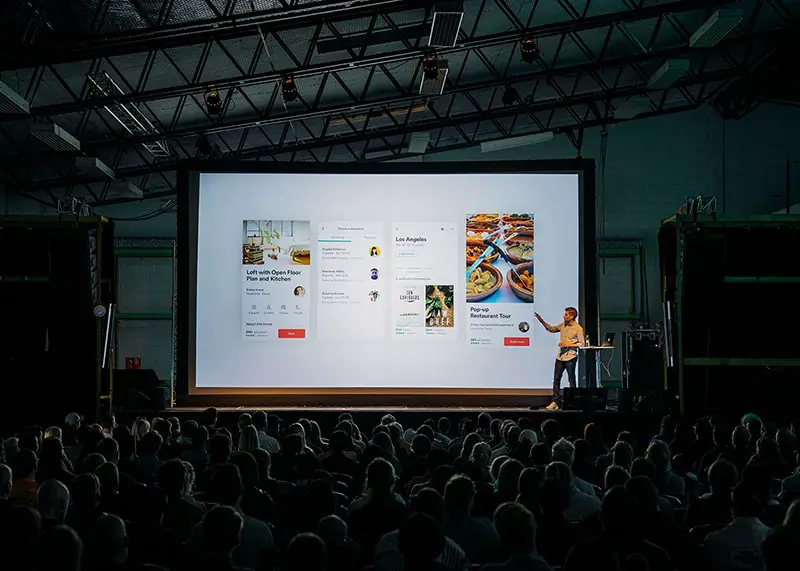 Person doing a business presentation standing in front of a large screen in front of people inside dim lit room