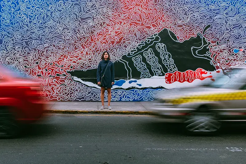 time lapse photography of woman standing on pavement in front of trainer artwork on wall