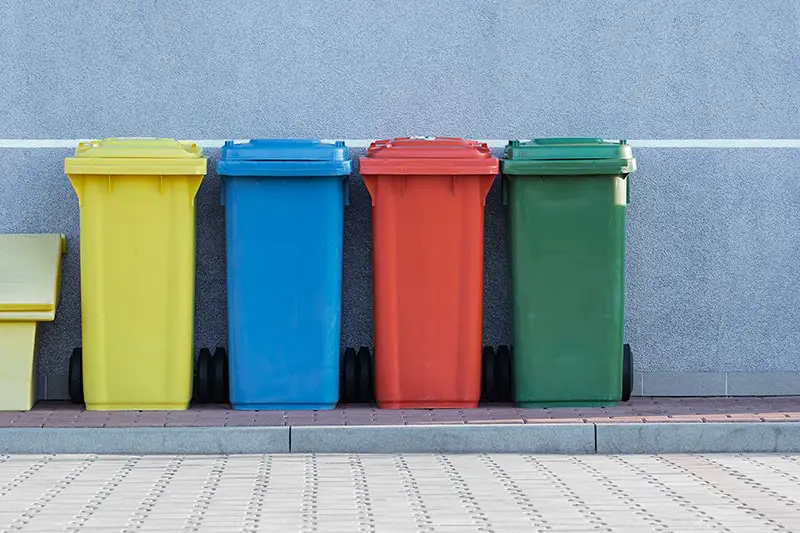 four assorted-color solid waste trash bins beside gray wall