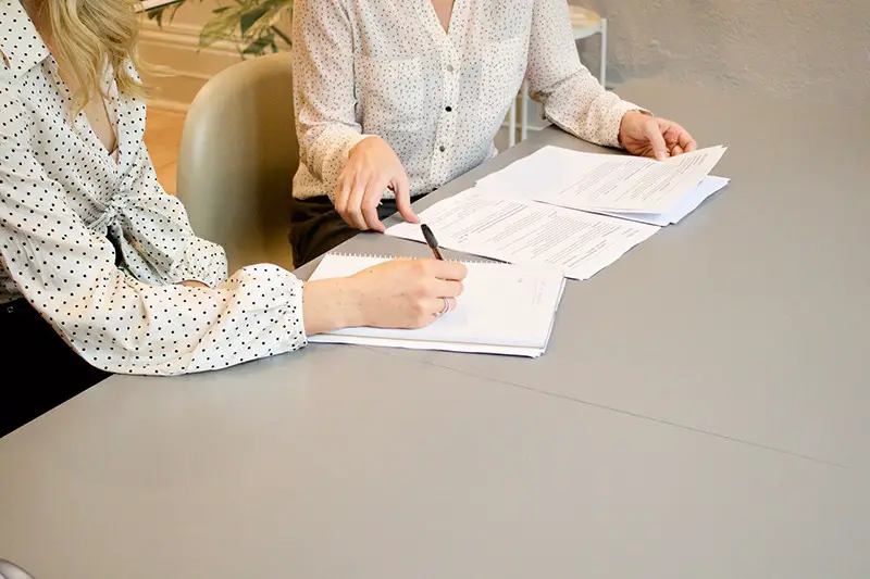 two women sat next to each other at a table reading and signing documents