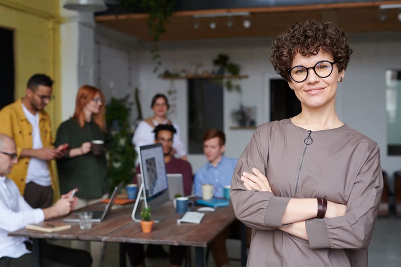 Woman standing in office wearing eyeglasses – with business colleagues behind her