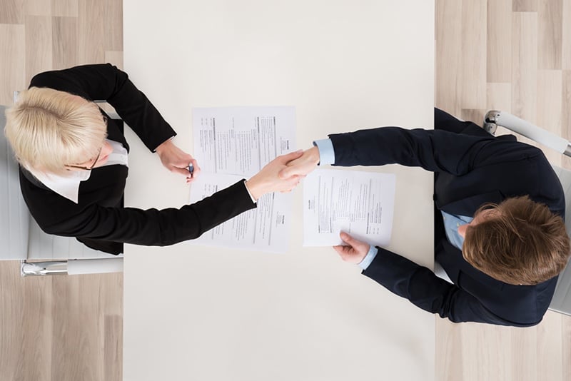 Two people shaking hands across a table and documents at a job interview