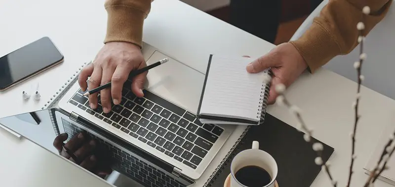 Person holding black pencil and notebook typing on laptop