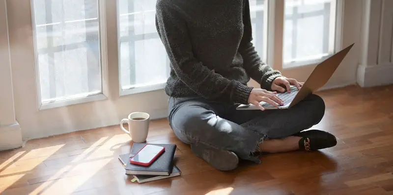 woman in grey sweater sitting cross legged on floor with laptop on knees