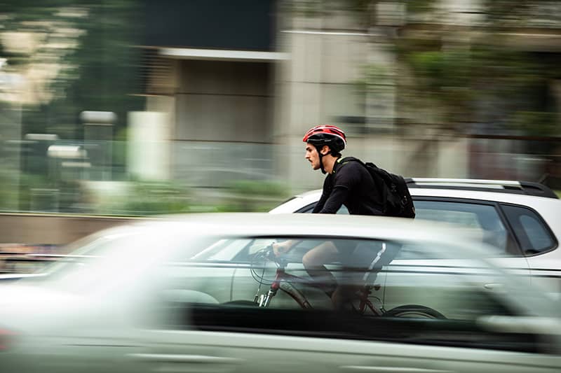 Man wearing a bike helmet and backpack on bicycle on road between cars
