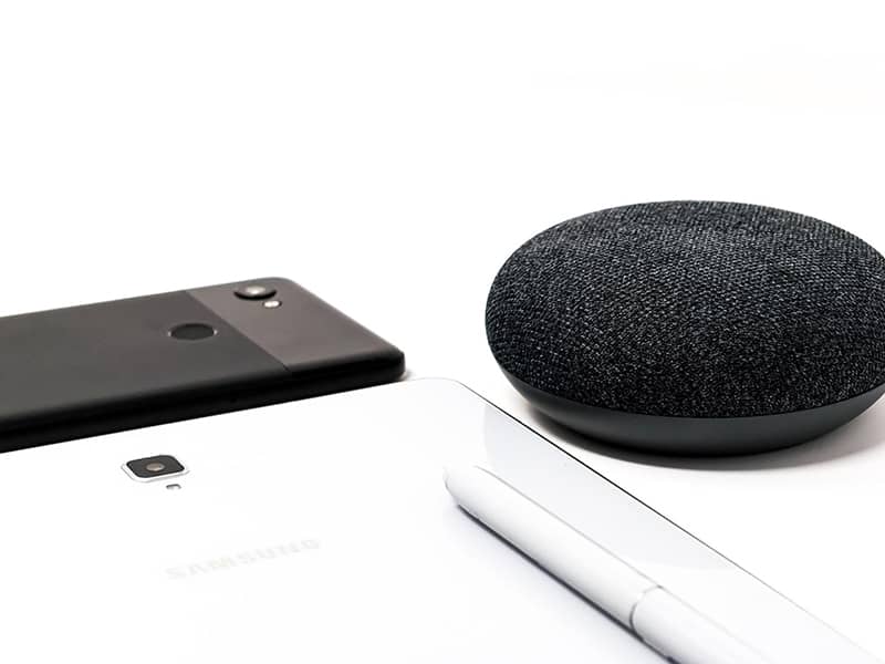 Black Phone, white tablet, white pen and Black Google home mini on a white table.
