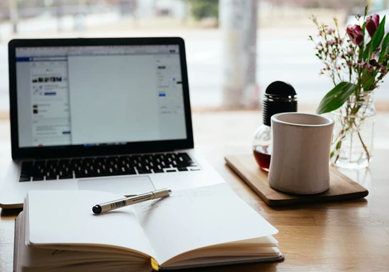 A laptop screen and the white cup and notebook in the table