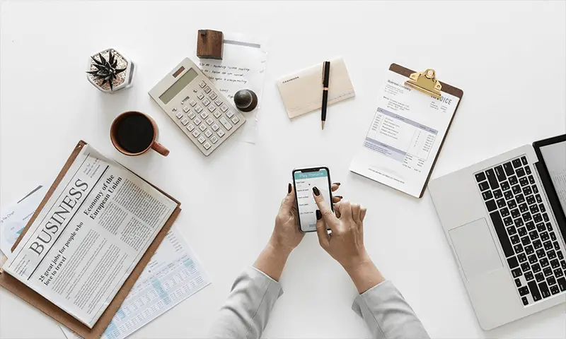 Person using smartphone at a desk, next to laptop, business newspaper, calculator and writing utensils