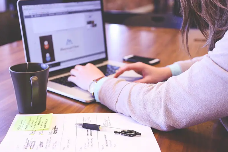 A brown mug, paper and pen, and a woman working on her laptop