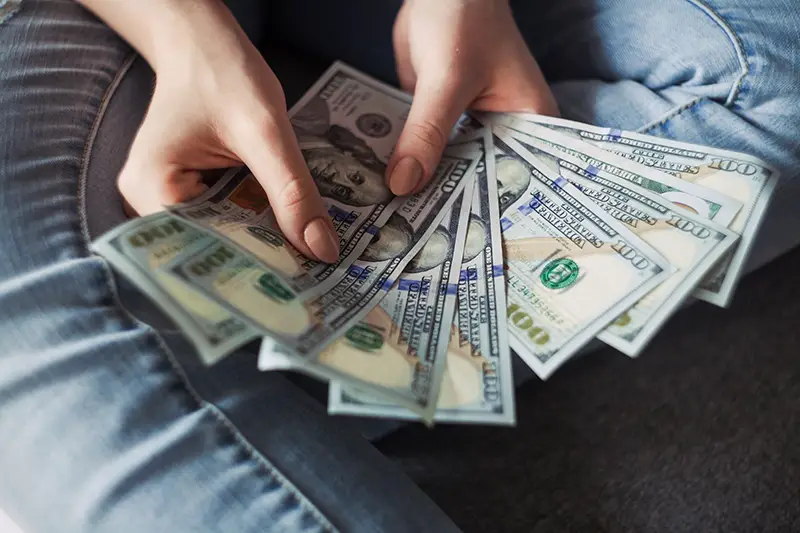 Person sitting on floor holding 100 dollar bills in a fan
