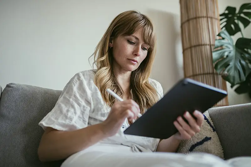 Woman using a stylus writing on a digital tablet while sitting in the couch