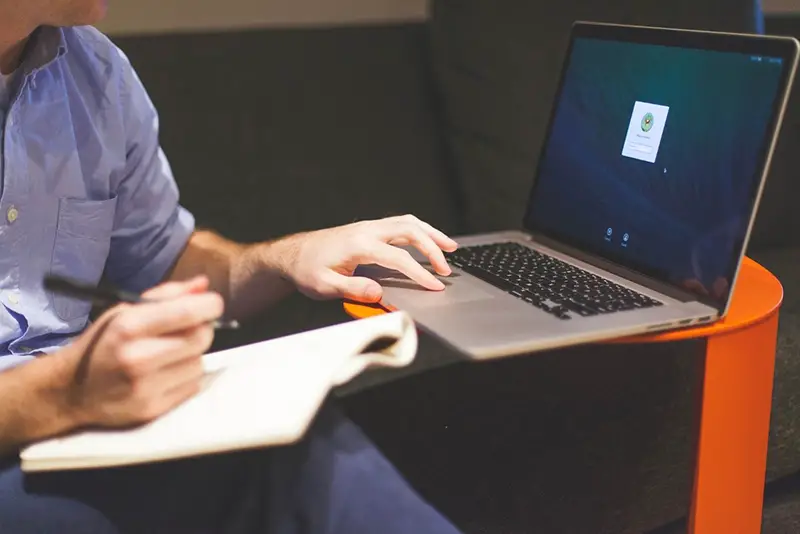 A person browsing in his laptop while holding pen and paper