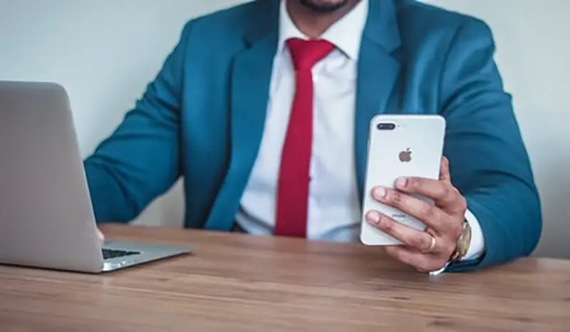 Man in blue suit holding an iPhone and typing on laptop computer