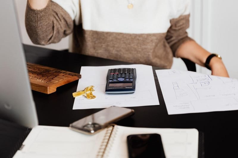 Woman at a desk next to a keyboard and monitor – calculator, paper, notebook and smart phones can be seen on the desk.