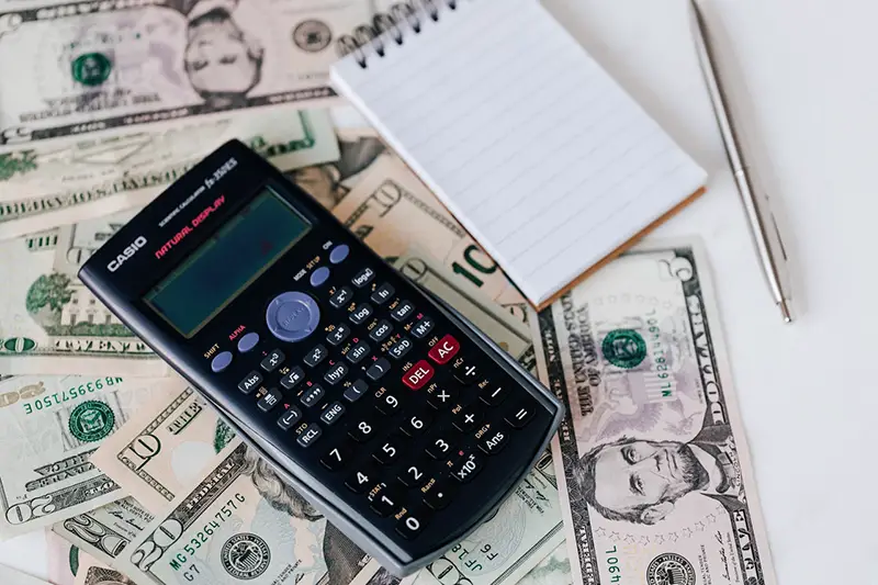 calculator, note pad and pen placed on stack of USA dollar bills