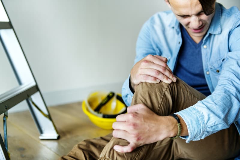 Employee with work injury - sitting on floor in pain holding leg, next to ladder and yellow hard hat