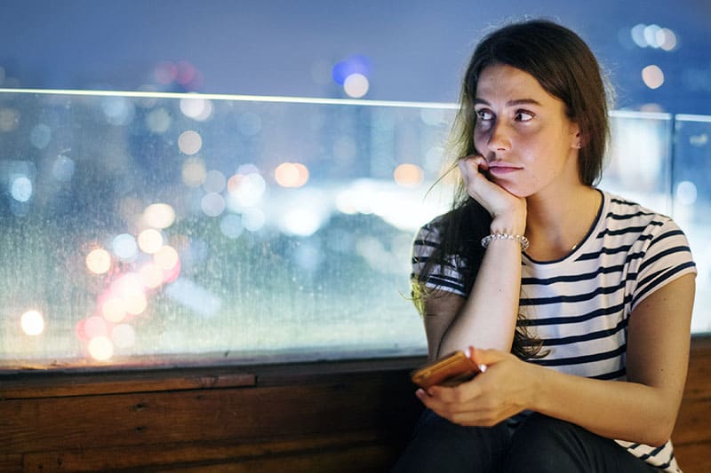 Unhappy young woman holding a smartphone in the evening cityscape