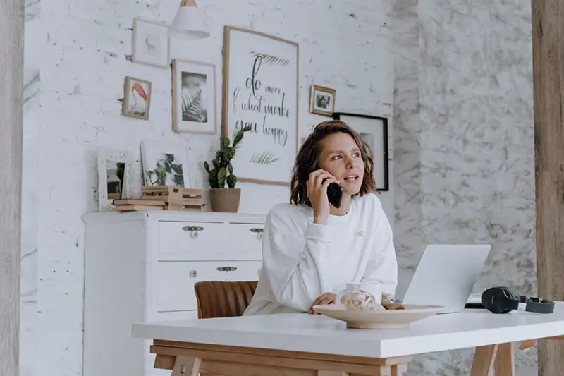 self-employed woman in white top speaking on the phone sat on wooden chair at her desk in front of her laptop