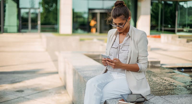 woman dressed in white and light coloured clothing sat on a wall texting on phone