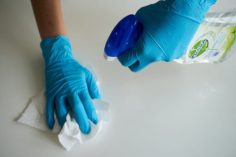 Person in blue gloves cleaning down a white counter top with Dettol spray and paper towel.