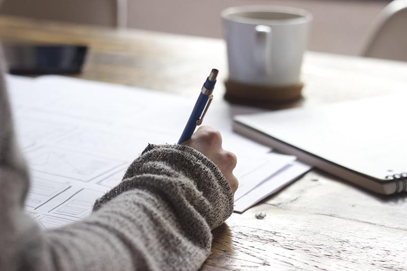 person writing on the paper with cup of coffee on the table