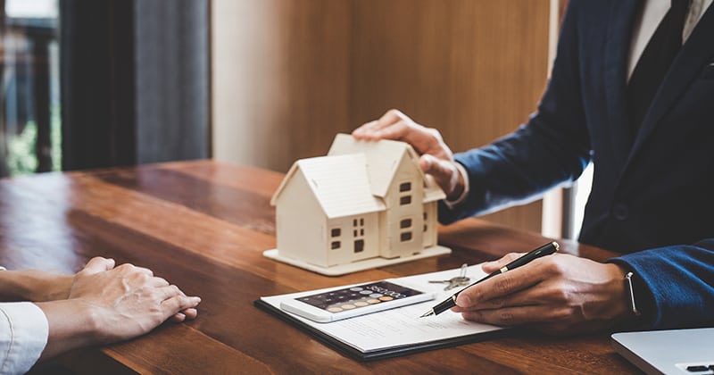 Two people in front of wooden table beside cartoon miniature house