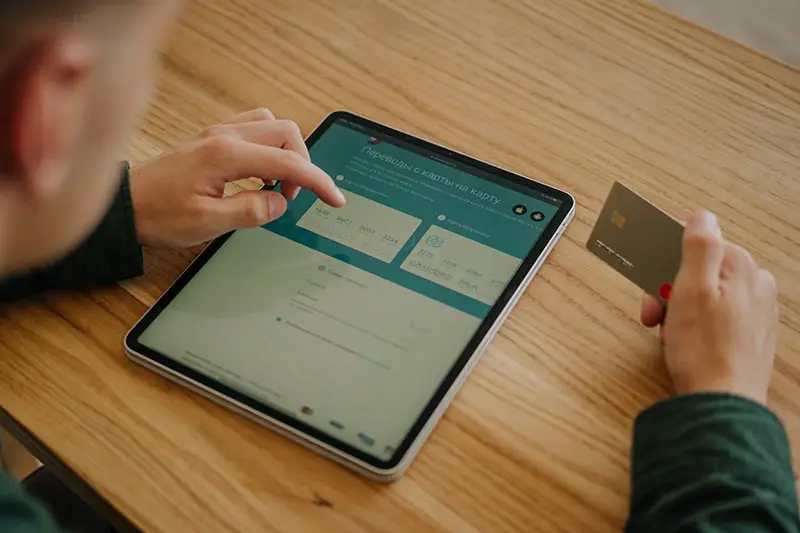 Person holding black Ipad and credit card on brown wooden table