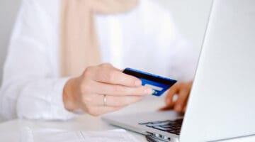 Person wearing white long sleeved shirt holding a credit card and making online payment on laptop
