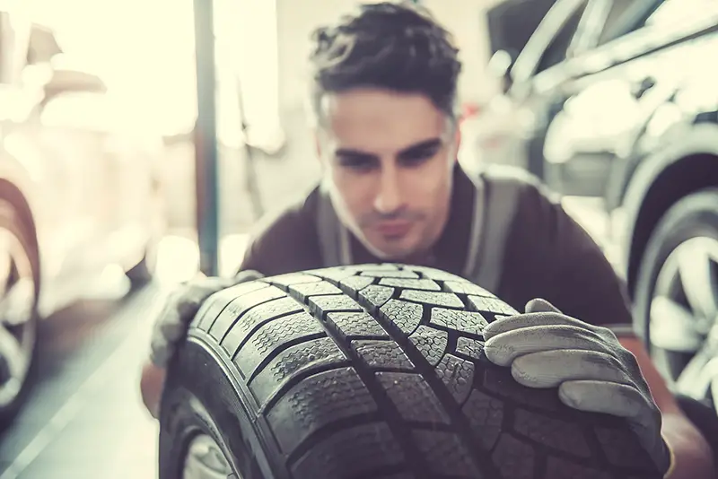 Handsome young auto mechanic in uniform is examining a tire while working in auto service