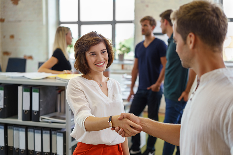 New hire - pretty young woman smiling while greeting man with handshake, standing in office environment with other people in background soft focus