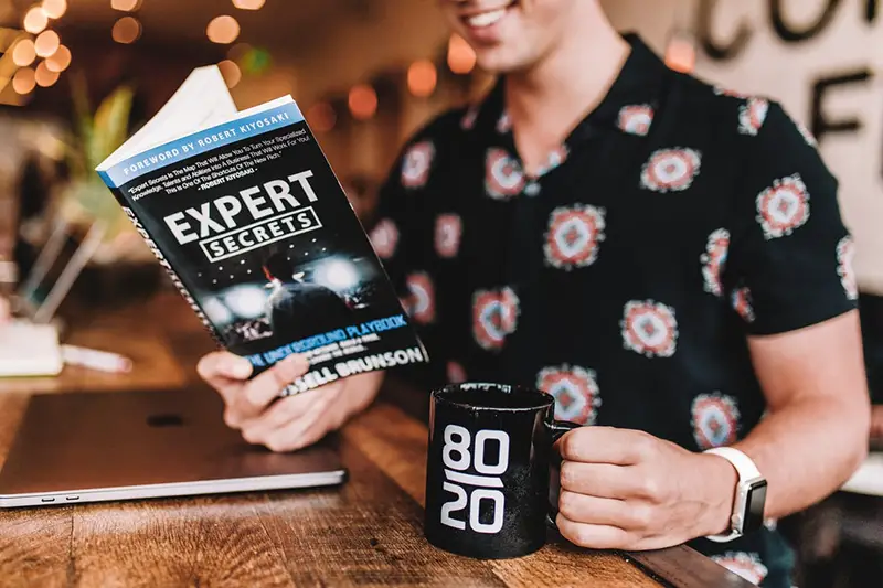 Person holding black mug while reading book of Expert secrets