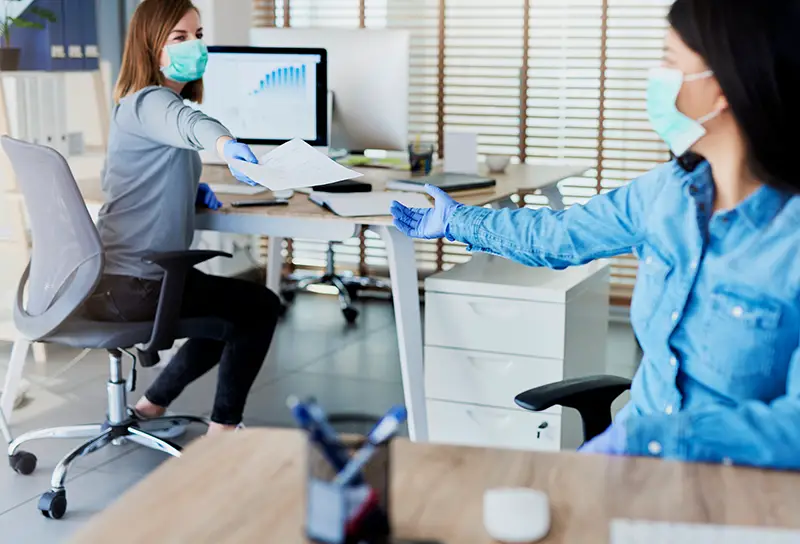 Two woman wearing facemask passing documents in the office