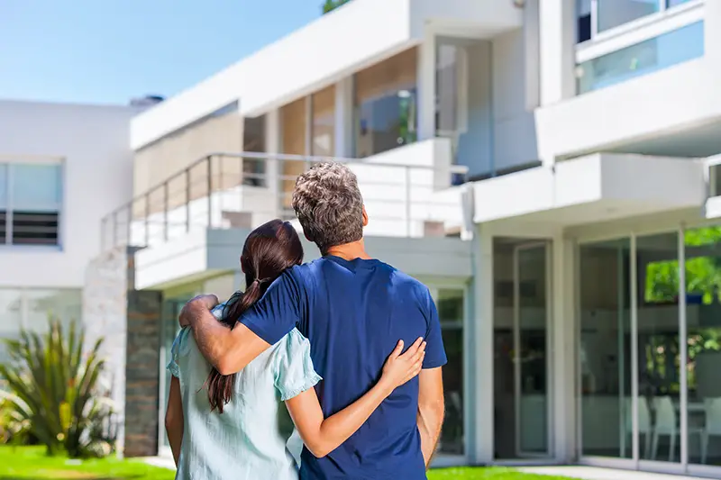 couple embracing in front of new big modern house, outdoor rear view back looking at their dream new home