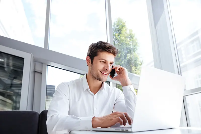 Happy young businessman talking on cell phone and using laptop in office