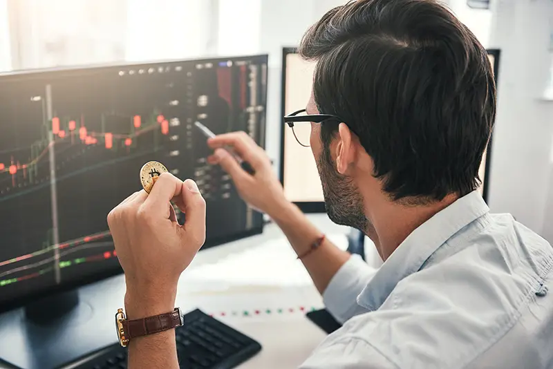 Young professional trader pointing on the data on computer screen with pen and holding Bitcoin in one hand