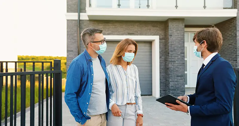 Caucasian young happy couple in medical masks buying house at outskirt and talking with male real-estate agent holding tablet device in hands.